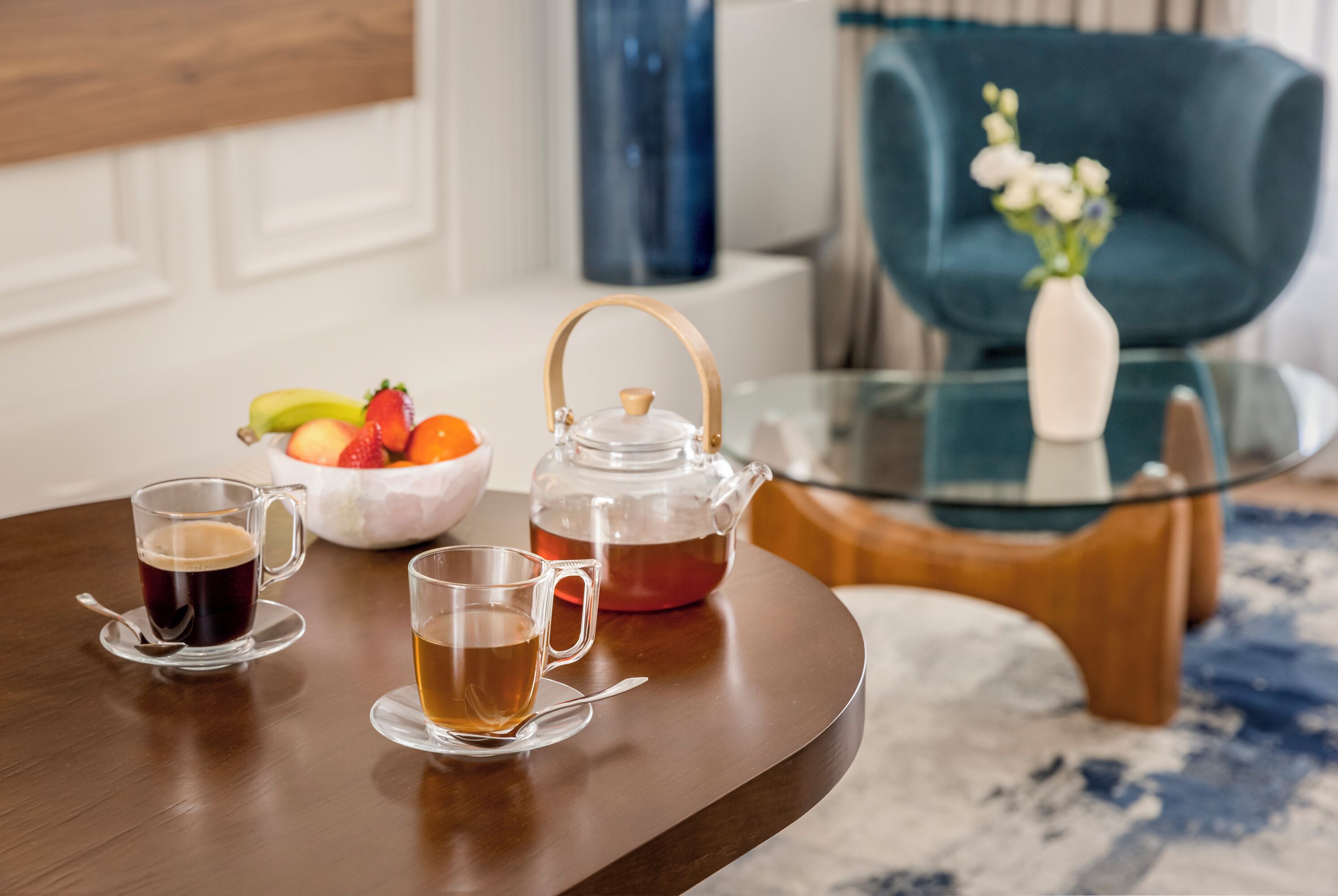 A wooden table with two cups of tea and a teapot, a bowl of fruit, and a vase with flowers on a glass table in a room with a blue chair and a white wall.