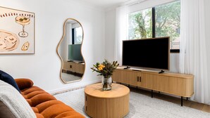 A modern living room with a sofa, a round wooden coffee table with a vase of flowers, a large flat-screen TV on a wooden stand, and a large mirror on the wall. - Bianca's- Stunning, Stylish & Steps from the Beach (Byron Bay)