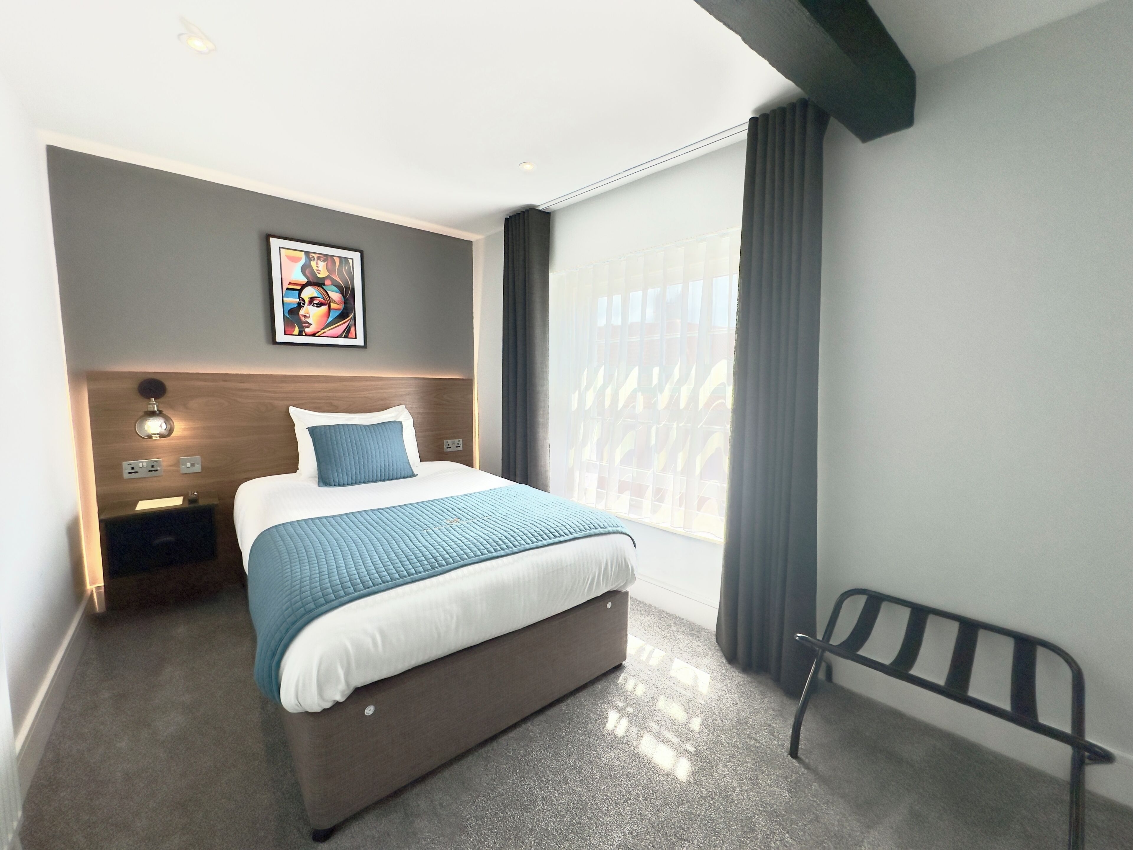 Una habitación de hotel moderna con una cama grande, cabecera con iluminación integrada, mesita de noche y ventana con cortinas. También se observa una obra de arte colgada en la pared.