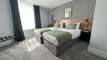 Quarto de hotel com duas camas, uma janela com cortinas, uma mesa de cabeceira e uma obra de arte na parede.