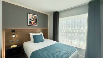 Una habitación de hotel con una cama, una mesita de noche, una lámpara de pared, una obra de arte enmarco y una ventana con cortinas.