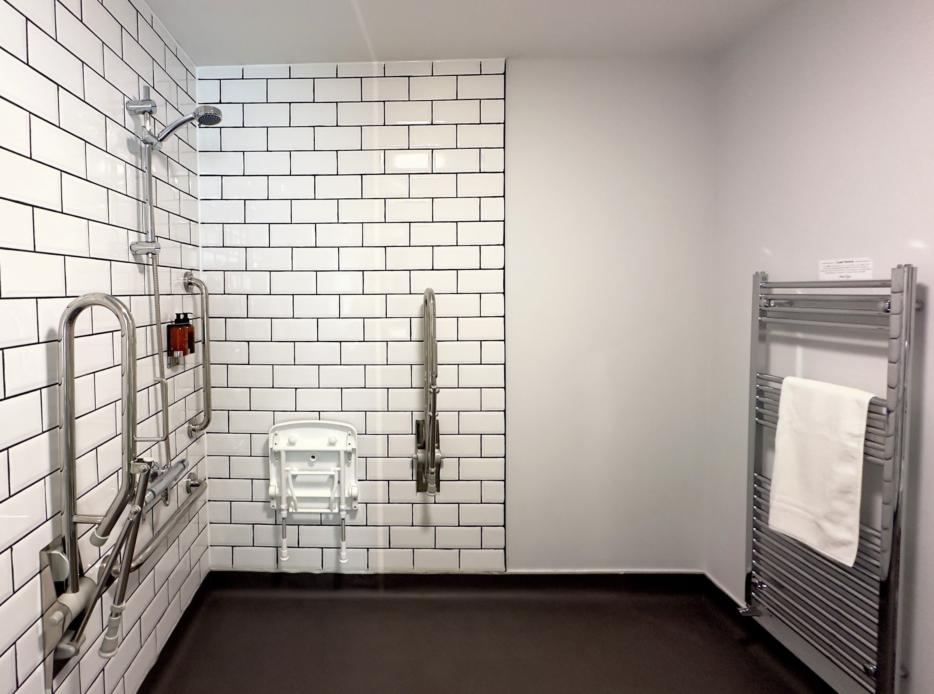 Un baño con azulejos blancos tipo metro, una ducha con cabezal de ducha manual, un perchero con una toalla y un secamanos.