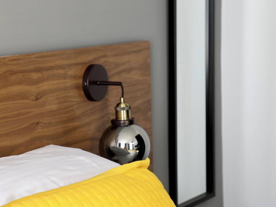 Een bed met een houten hoofdbord, een bedlampje en een grote spiegel aan de muur.