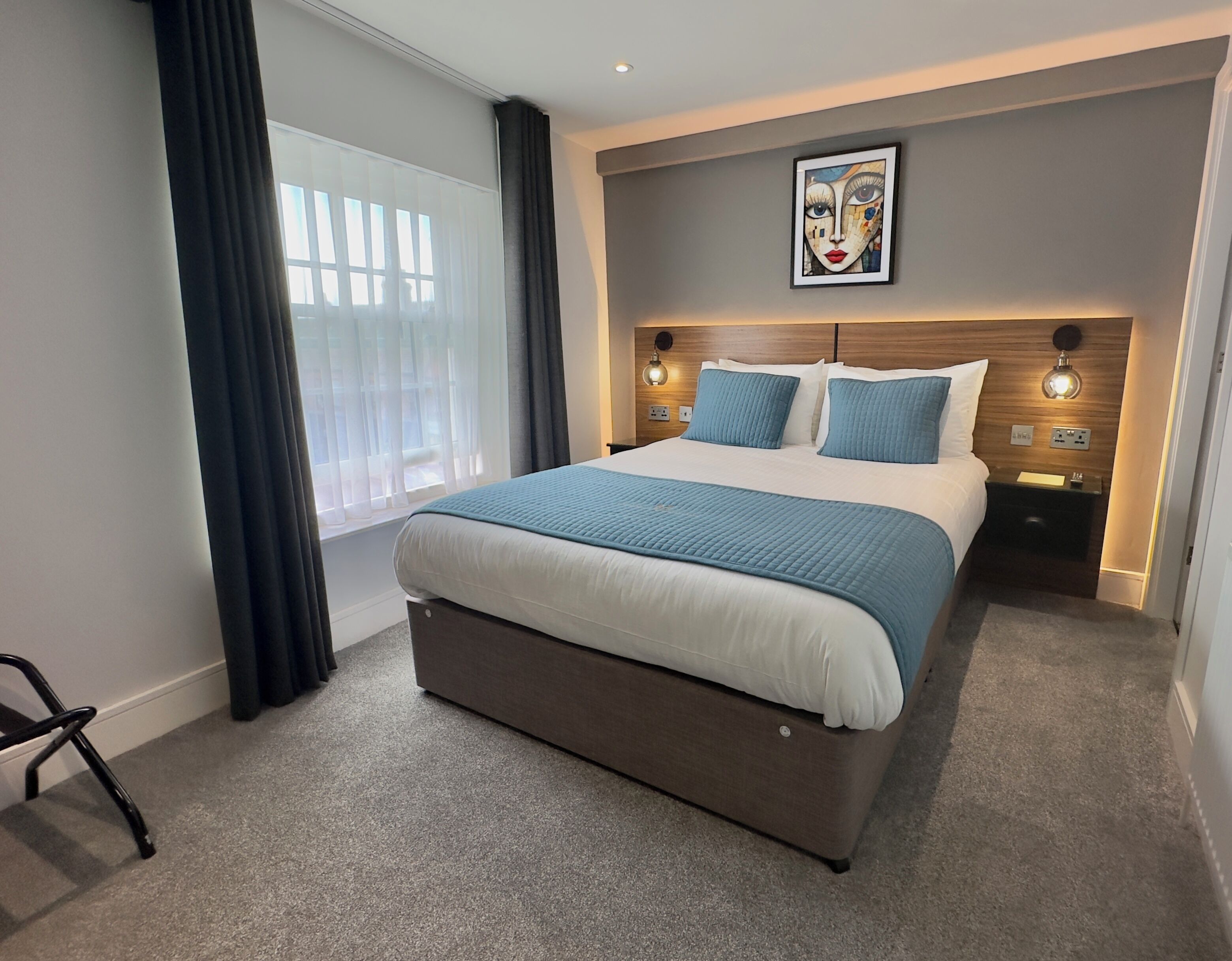 A modern hotel room with a large bed, a headboard with integrated lighting, a bedside table with a lamp, and a framed artwork on the wall.