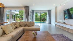 Ein modernes Wohnzimmer mit einer Eckcouch, einem Couchtisch mit floralem Mittelstück, einem wandmontierten Flachbildfernseher und großen Fenstern mit Blick ins Freie.
