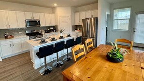 A modern kitchen with white cabinets, stainless steel appliances, and a wooden dining table with bar stools. - Spacious 4BD3BA, Game Room & 2 King Beds & Patio (Austin)