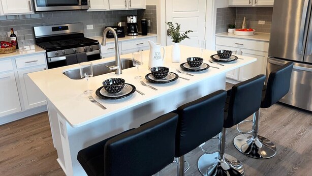 A modern kitchen with white cabinets, a stainless steel refrigerator, a microwave, a stove, and a sink. There are four black bar stools around the island. - Spacious 4BD3BA, Game Room & 2 King Beds & Patio (Austin)
