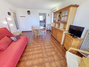 Un salon avec un canapé rouge, une table à manger avec des chaises, un coin cuisine avec des meubles en bois et un téléviseur posé sur un meuble en bois.