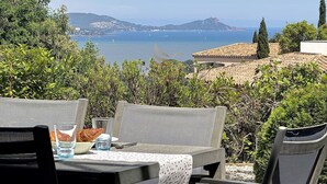 Een terras met een tafel voor twee, met uitzicht op de kust, boten en heuvels in de verte.