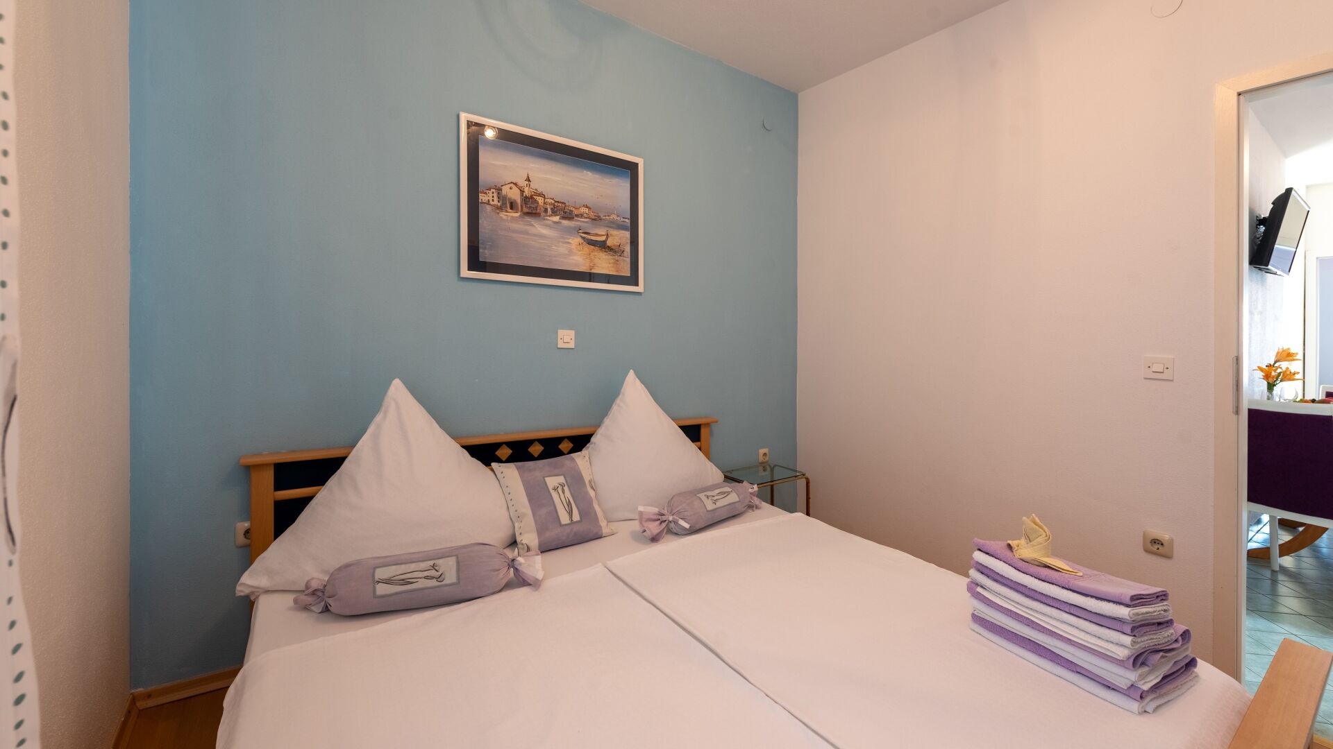 Ein Schlafzimmer mit einem Bett, Kissen, einem Nachttisch, einem gerahmten Bild an der Wand und einem Stapel Handtücher.