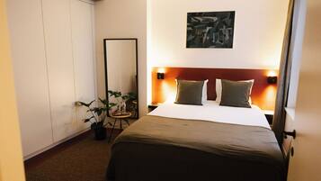 A hotel room with a large bed, a bedside table, a mirror, a wall-mounted artwork, and a potted plant.