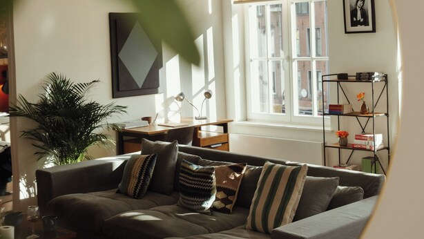 Een gezellige woonkamer met een hoekbank, een salontafel, een boekenkast en een groot raam dat voor natuurlijk licht zorgt.