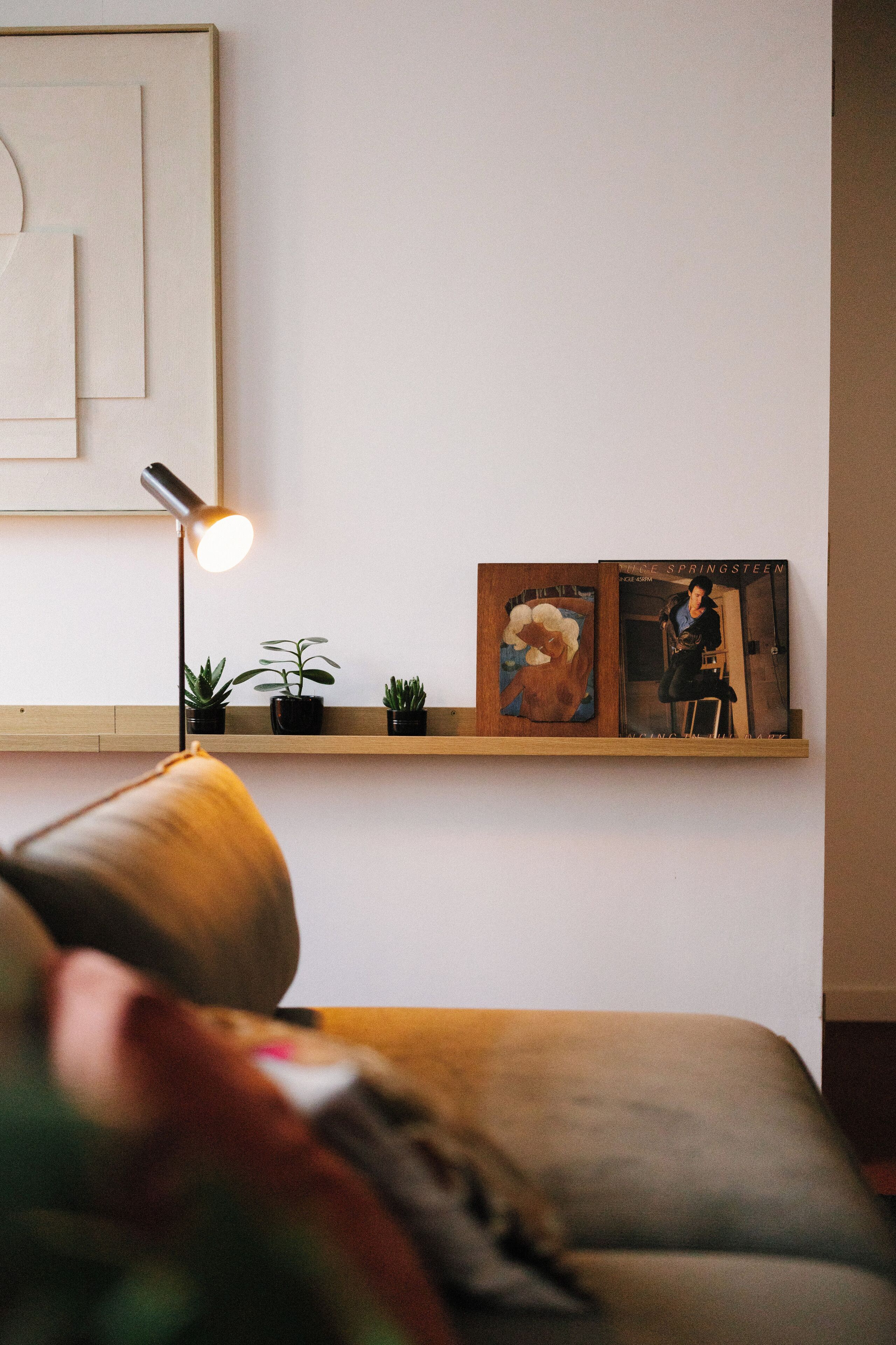 A minimalist room with a bed, a bedside lamp, and a shelf with books and small plants.