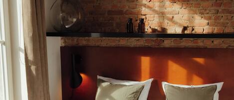 A bedroom with a brick wall, a bed with white bedding, and a shelf above the bed.