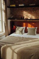 A bedroom with a brick wall, a bed with white bedding, and a shelf above the bed.