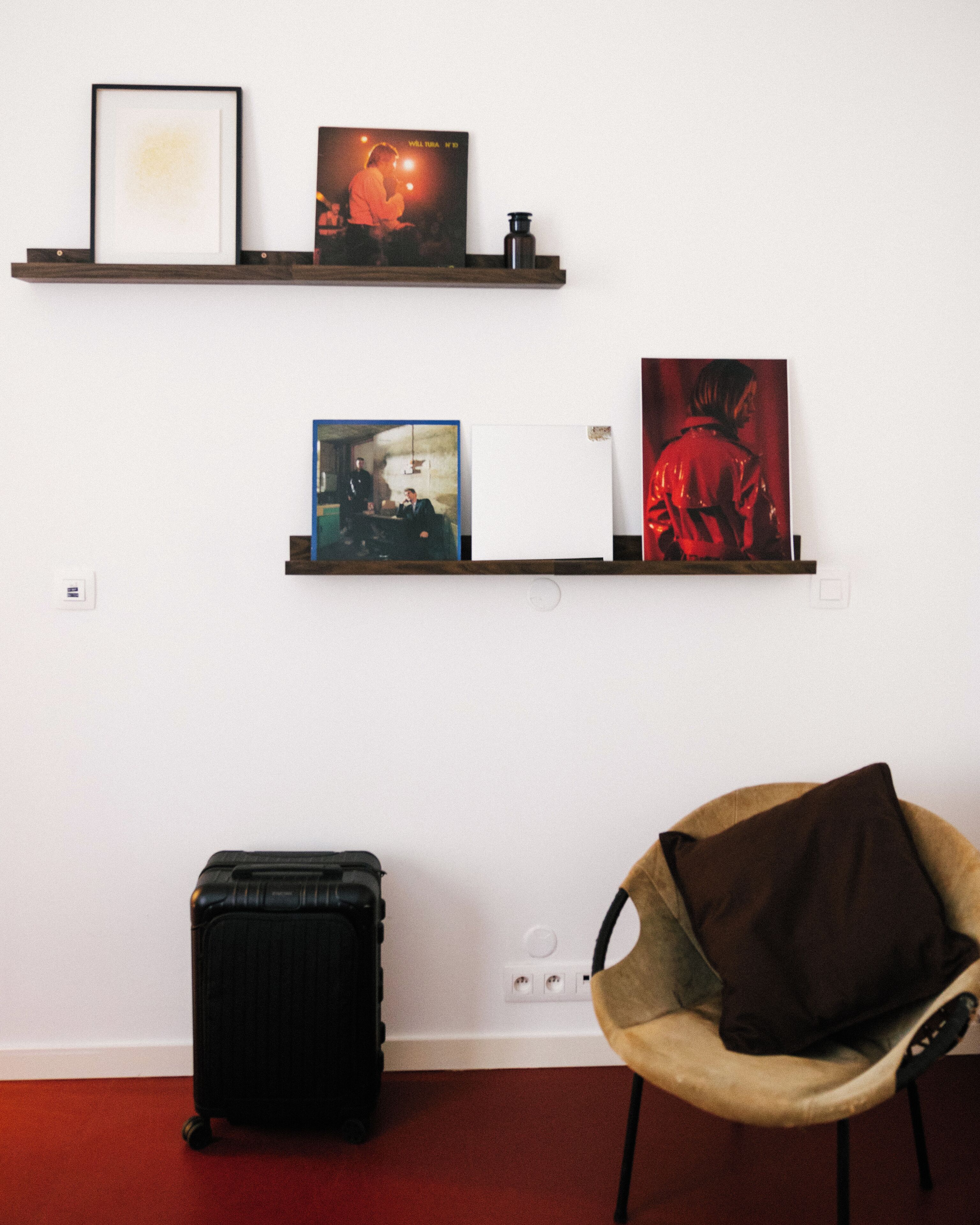 Una habitación con dos estantes empotrados que exhiben obras de arte enmarcadas, una maleta negra y un sillón con cojín.