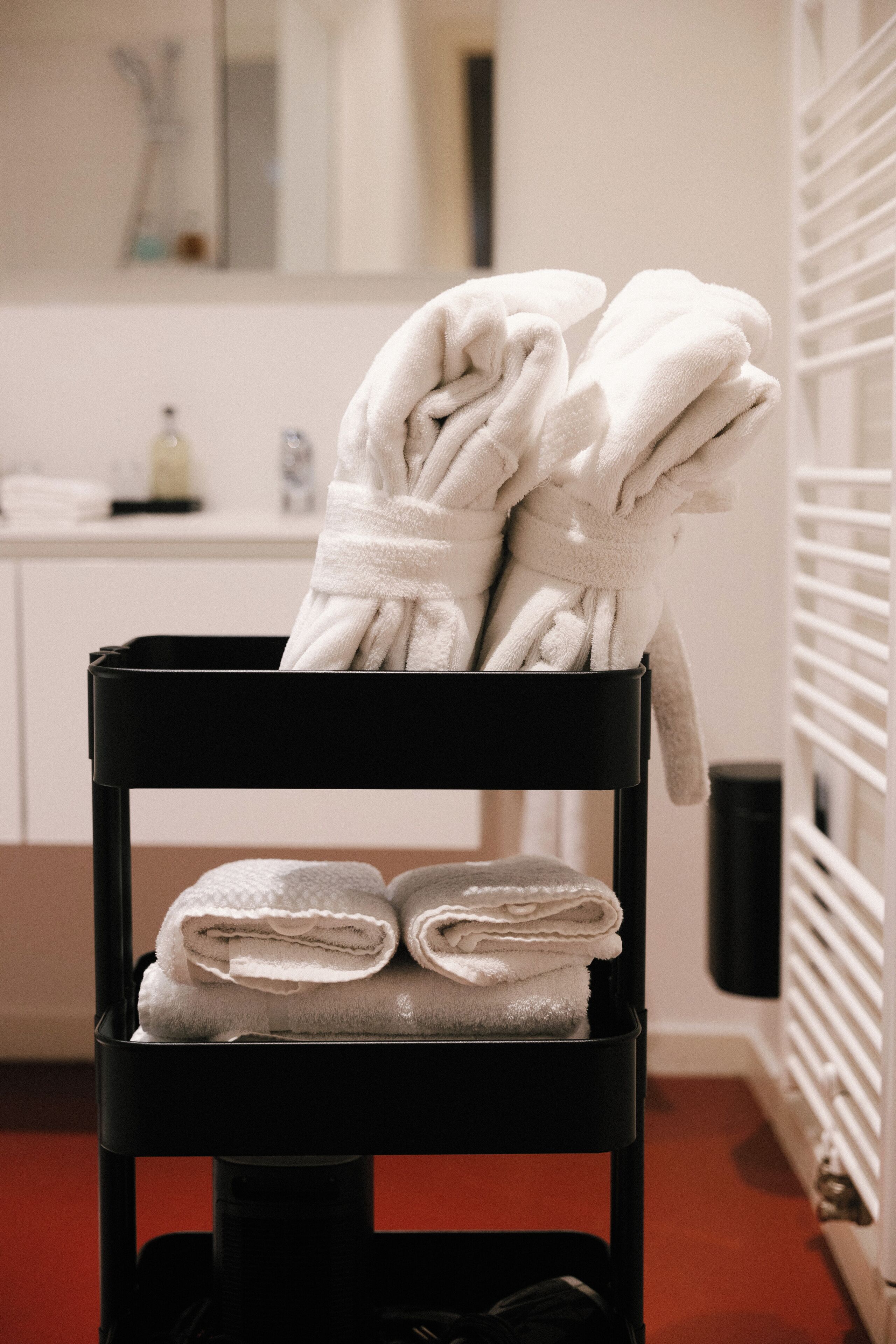 Una silla negra con una pila de toallas blancas, ubicada en un baño con un lavabo blanco y un radiador al fondo.