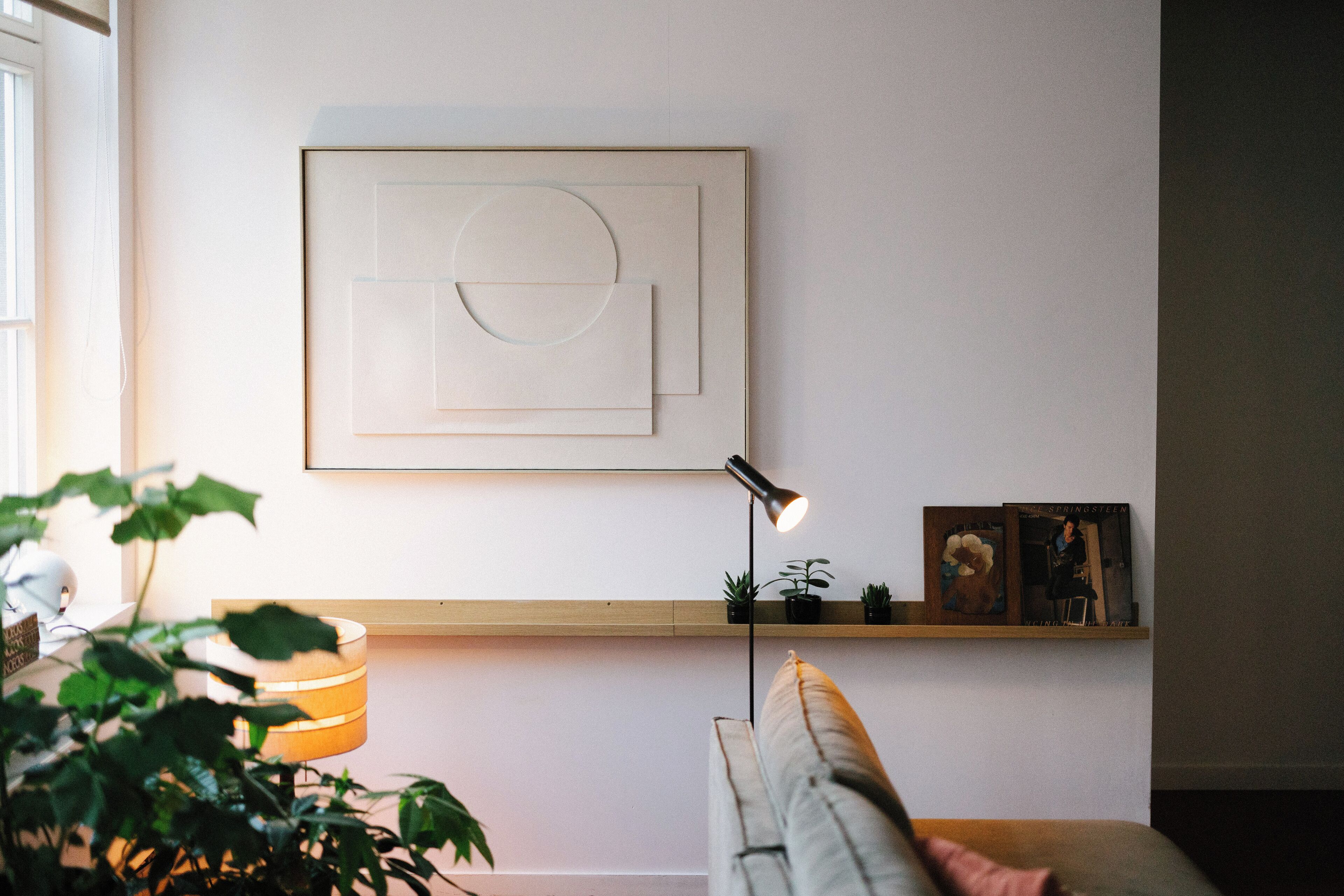 A minimalist living room with a wall-mounted abstract artwork, a floor lamp, a bookshelf with books and plants, and a couch.
