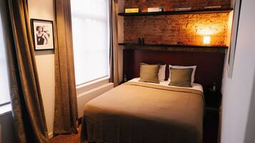 A bedroom with a brick wall, a bed with a beige bedspread, two pillows, a nightstand, and a shelf with books and a lamp.