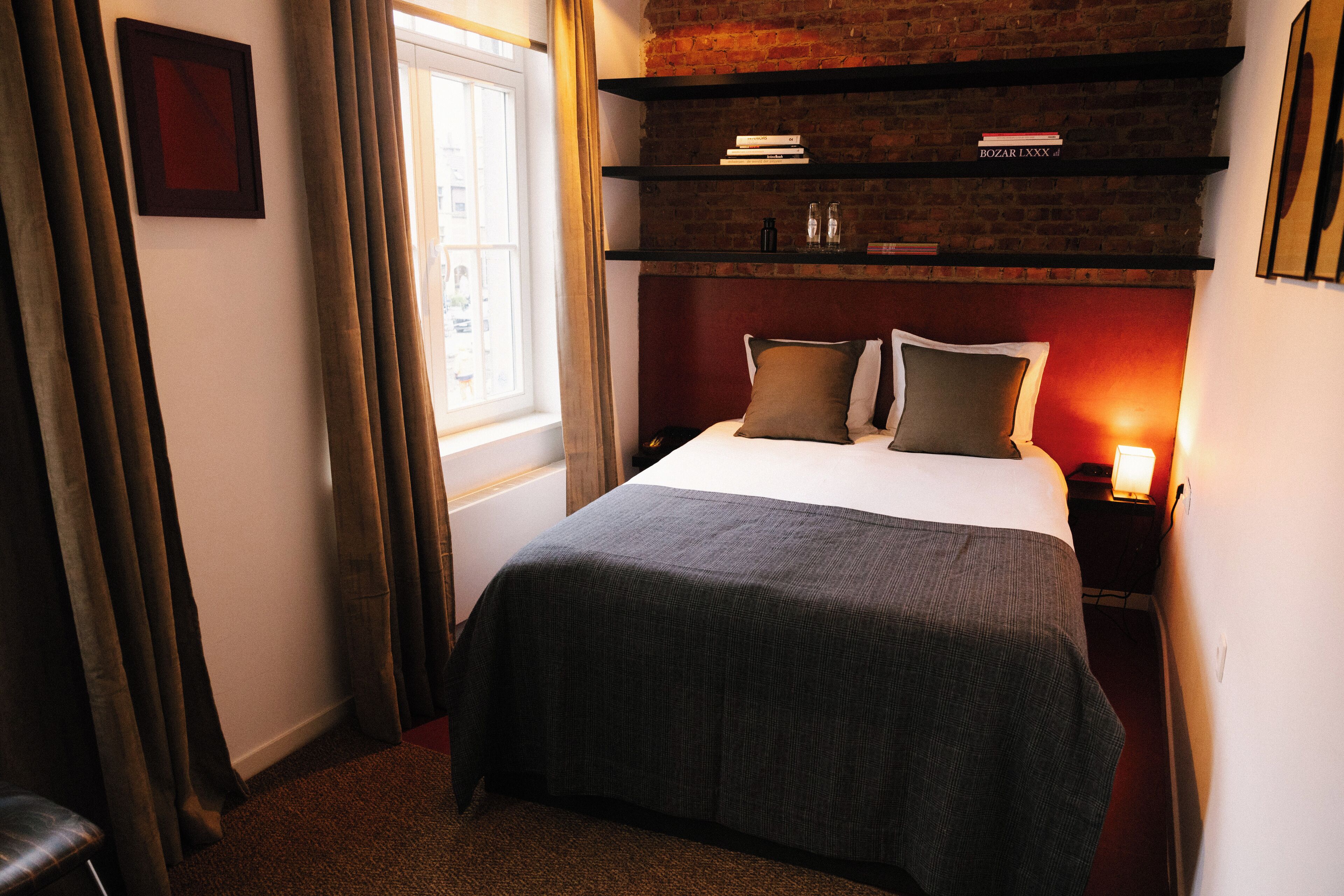Habitación de hotel con cama, mesita de noche con lámpara, ventana con cortinas y pared de ladrillo.