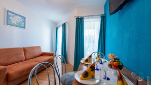 Een kamer met een bruine bank, een eettafel gedekt met borden, glazen en fruit, en een aan de muur bevestigde televisie.