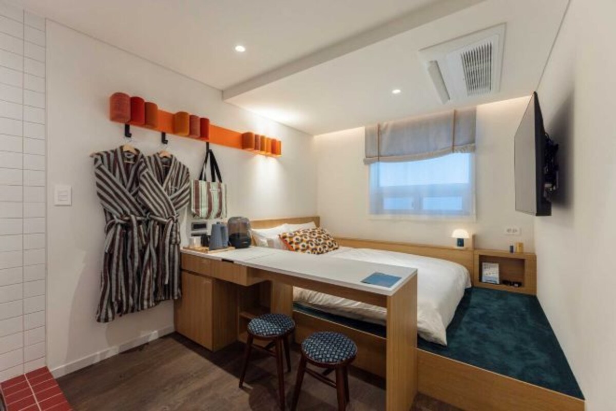 A compact hotel room with a bed, a desk with a chair, a small seating area, and a TV mounted on the wall.