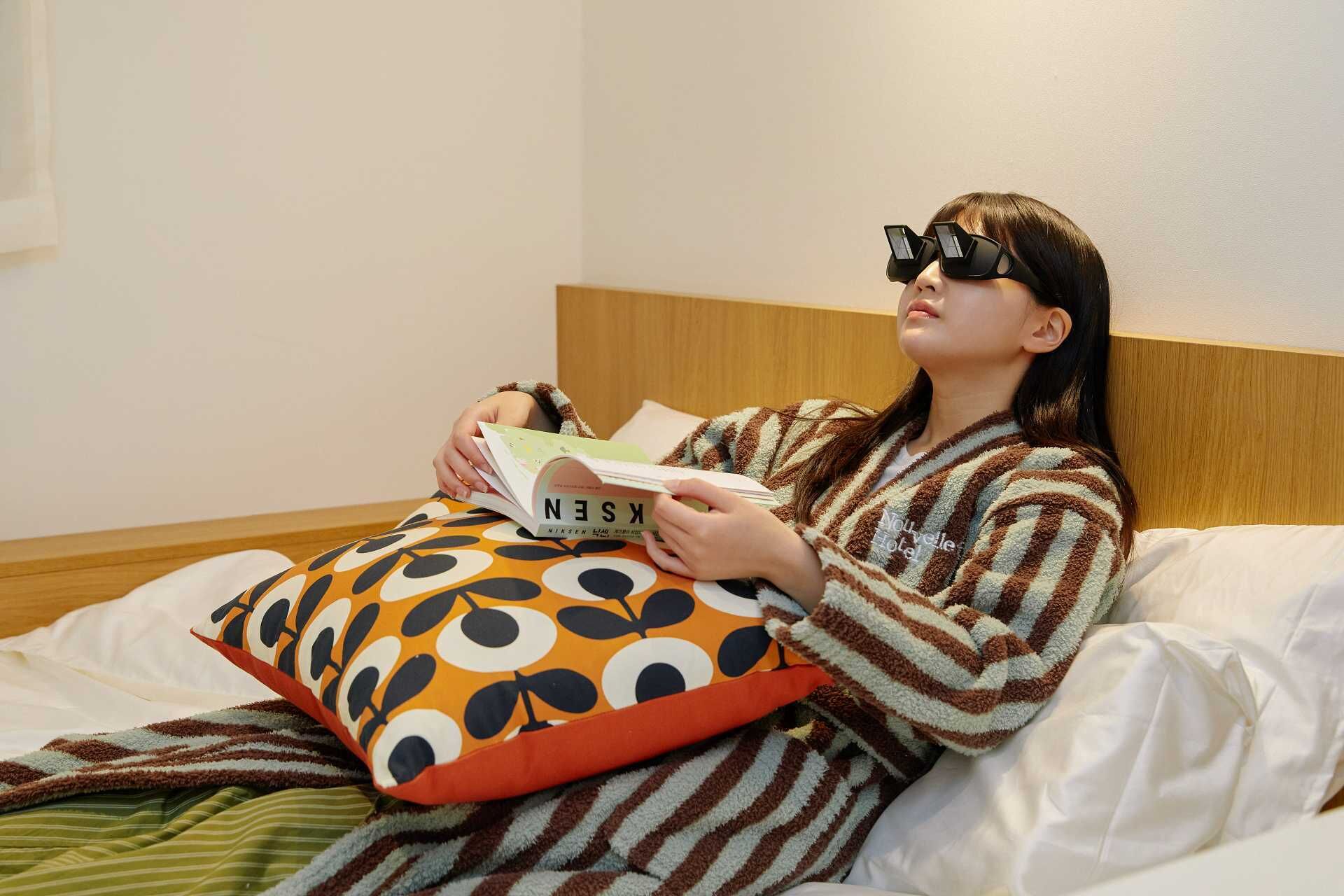 Una persona sentada en una cama leyendo un libro, vestida con una bata a rayas y usando gafas 3D.