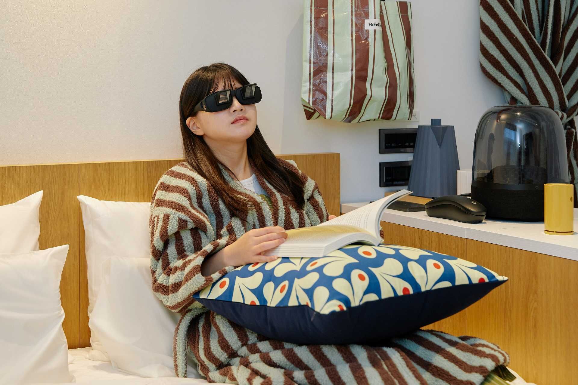 A person sitting on a bed with striped bedding, reading a book. There is a pillow with a blue and white pattern, a black telephone, and a yellow object on the bedside table.