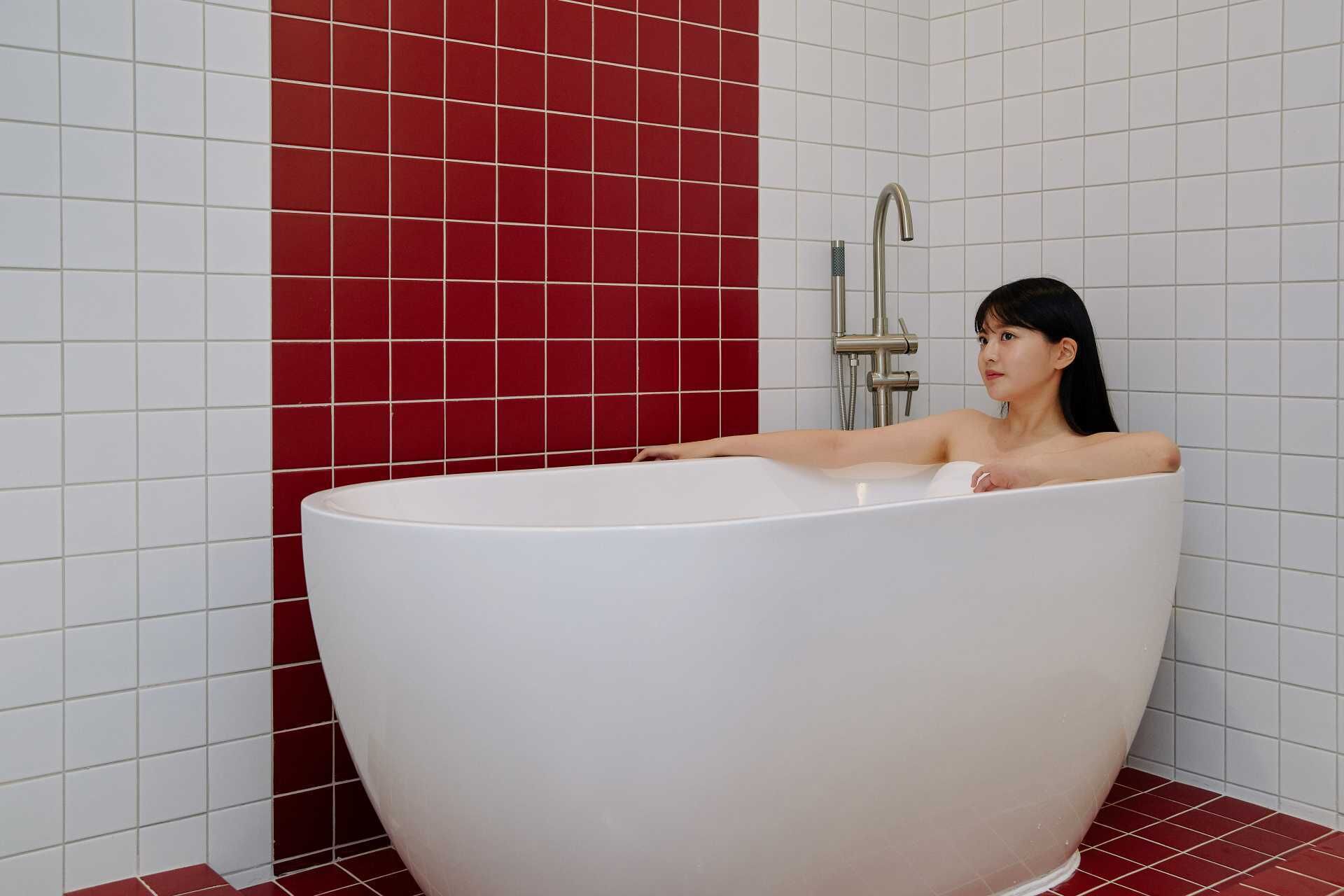 A person in a large, white freestanding bathtub in a bathroom with red and white tiled walls.