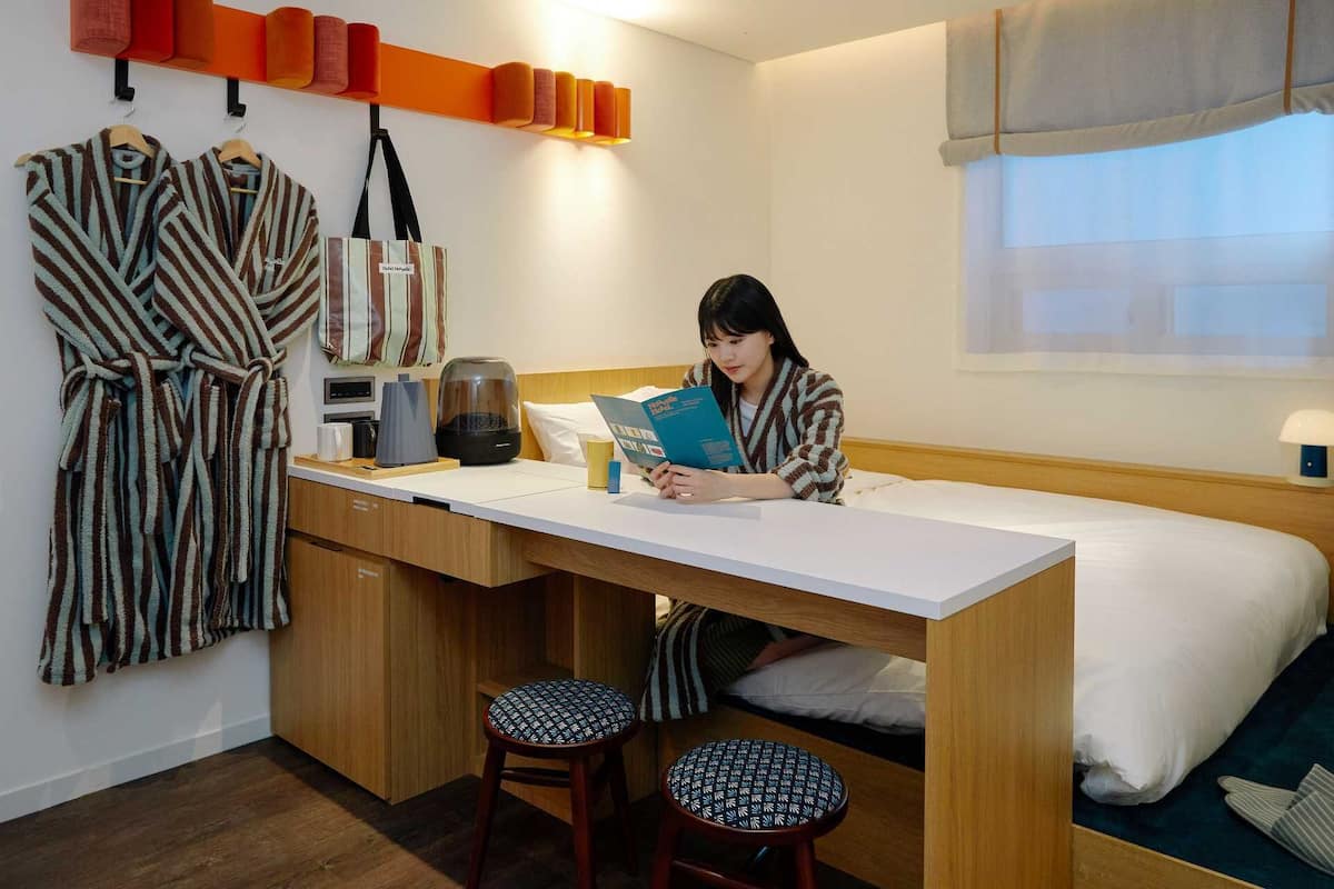 Habitación de hotel pequeña con cama, escritorio, dos taburetes, cafetera y bata colgada en la pared.