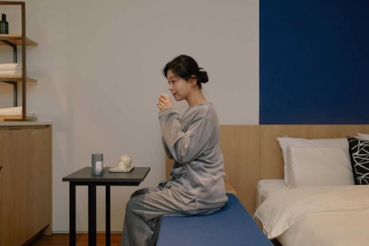 A person sitting on a bed in a room with a small table, a shelf with items, and a bed with pillows.