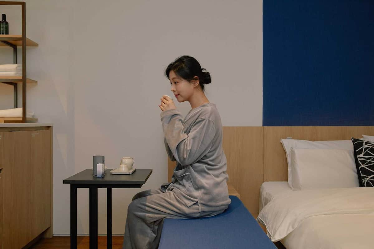 Une personne est assise sur un lit dans une pièce où se trouvent une petite table, une étagère avec des serviettes et un mur bleu.