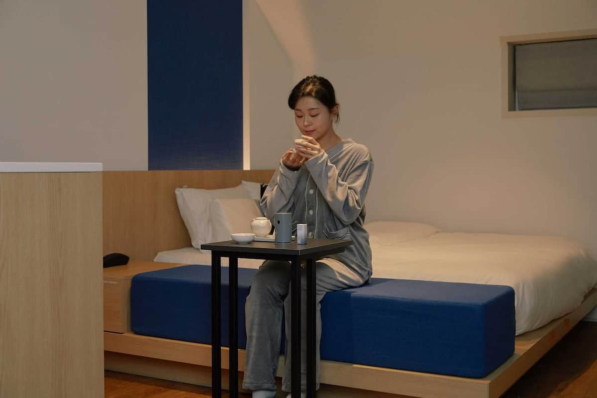 Une personne est assise sur un lit dans une pièce où se trouve une petite table.