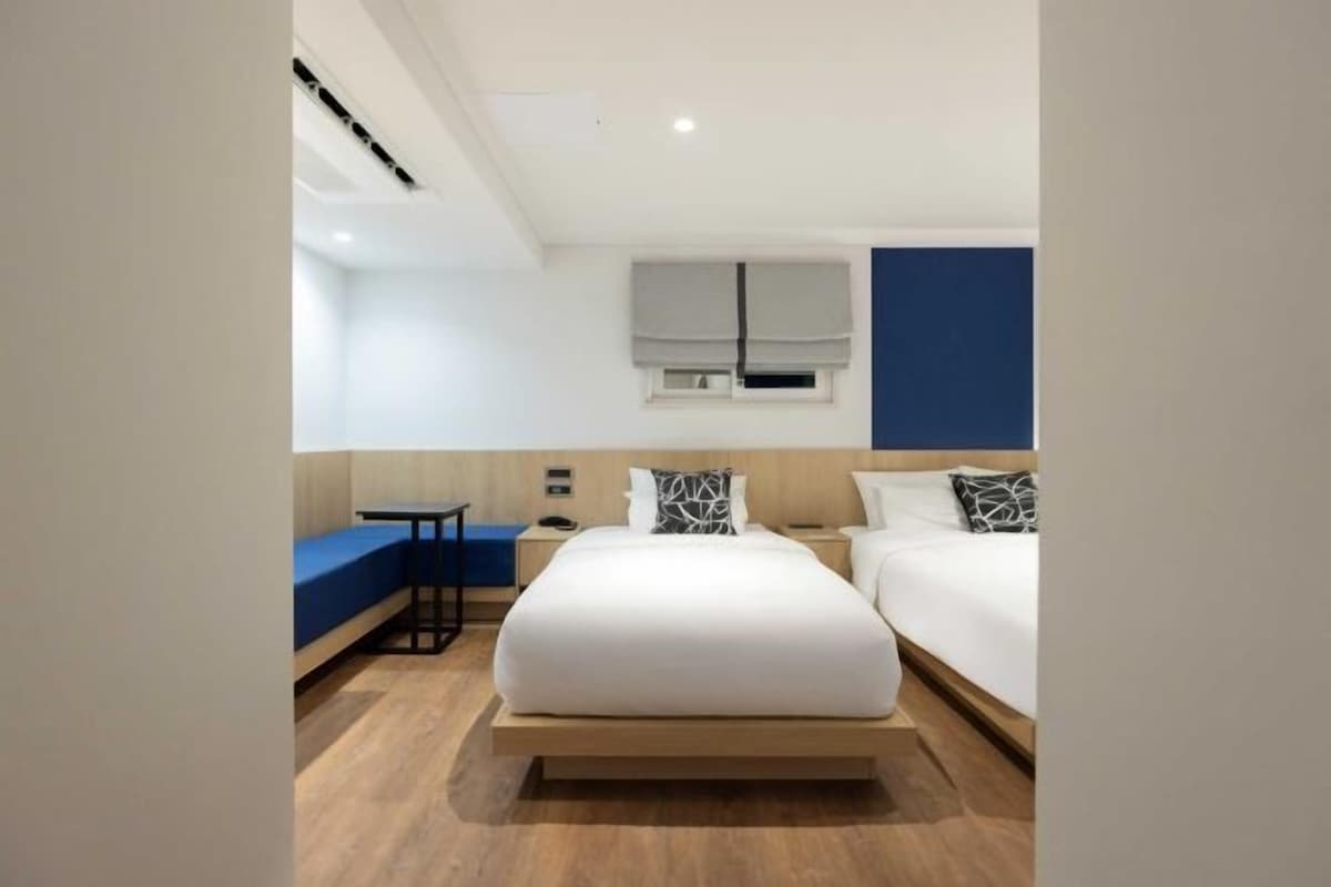 A modern hotel room with a large bed, a bedside table, a small desk, and a blue bench.