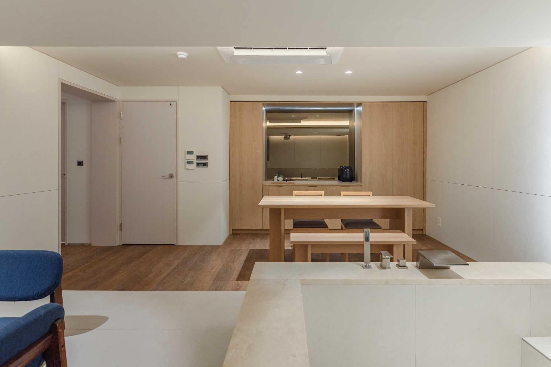 A modern, minimalist room with a wooden dining table, a bench, and a small kitchenette.