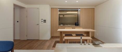 A modern, minimalist room with a wooden dining table, a bench, and a small kitchenette.