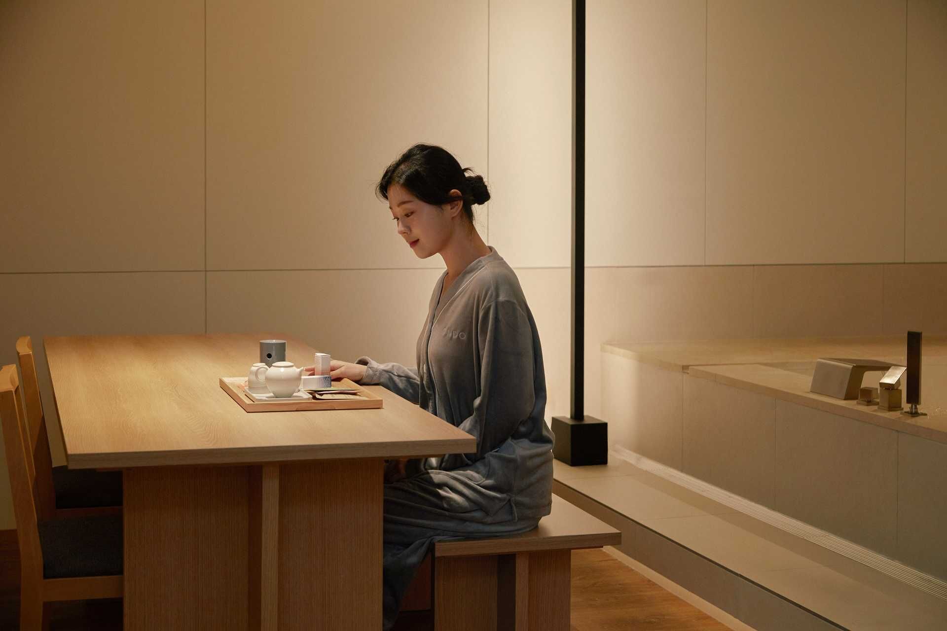Une personne est assise à une table en bois, devant une théière et des tasses, dans une pièce au design minimaliste, éclairée par un grand lampadaire.