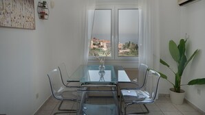 Un comedor con una mesa y sillas de vidrio, una planta en maceta y vistas de edificios a través de una ventana.