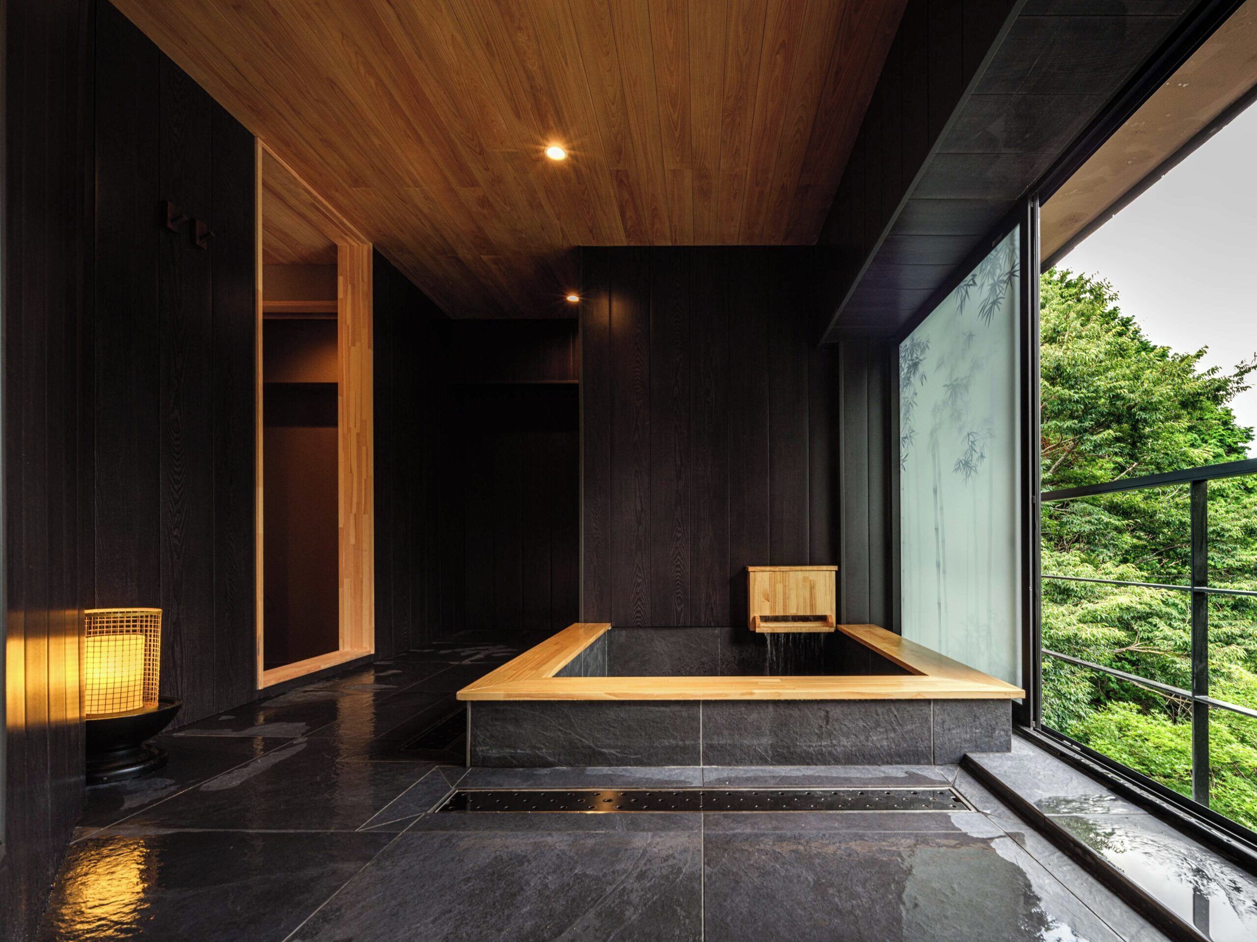 A modern interior with a large bathtub, dark wooden walls, and a view of greenery outside.