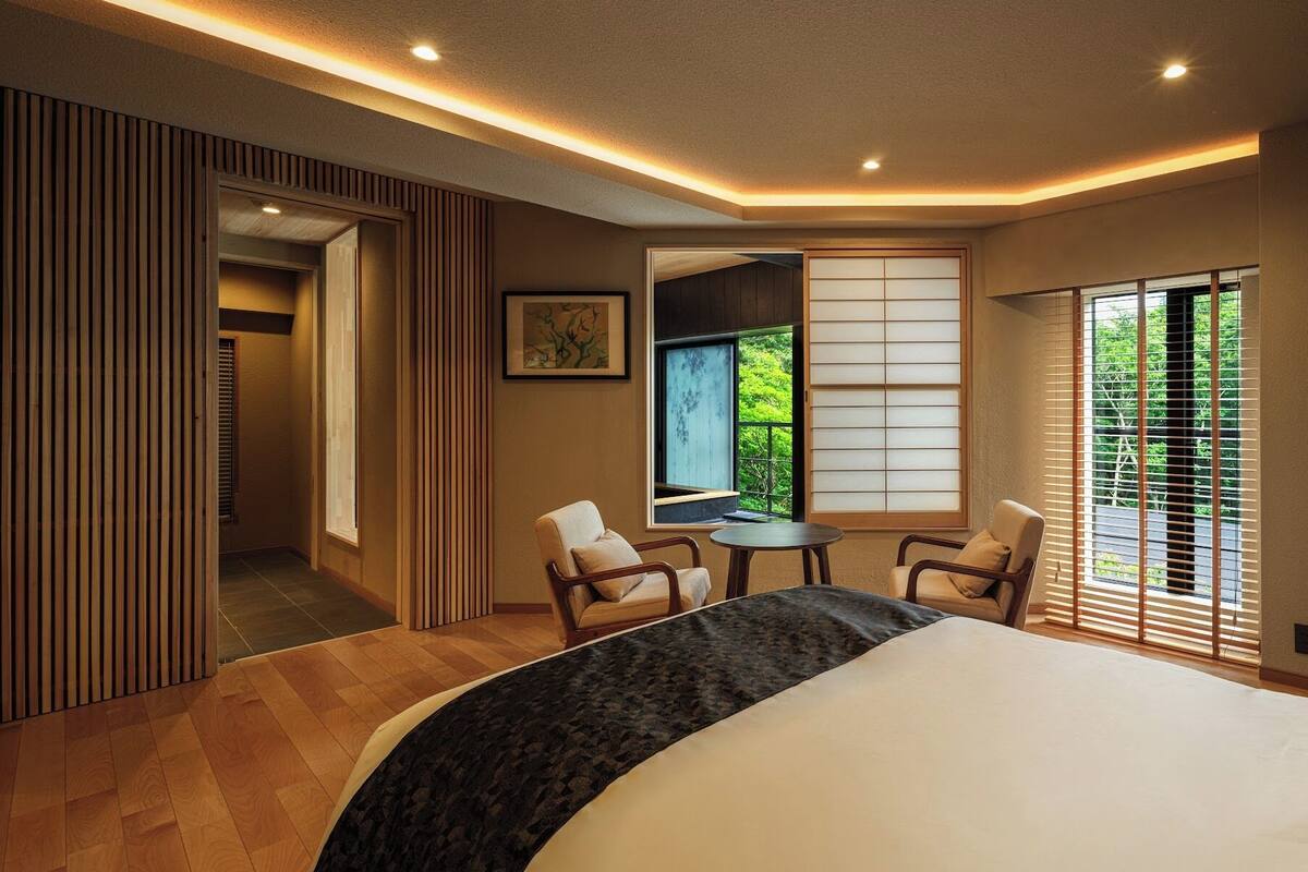 Een moderne slaapkamer met een groot bed, een klein tafeltje, twee stoelen, een schuifdeur en uitzicht op groen.