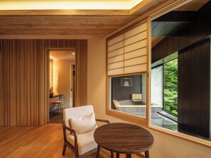 A modern interior with a wooden floor, a round table, a cushioned chair, and a large window with a view of greenery. - Hakonehibarino (Hakone)