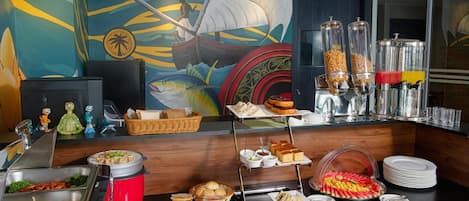 Een buffetopstelling met diverse gerechten, waaronder fruit, belegde broodjes en drankjes, plus een muurschildering van een zeilscene.