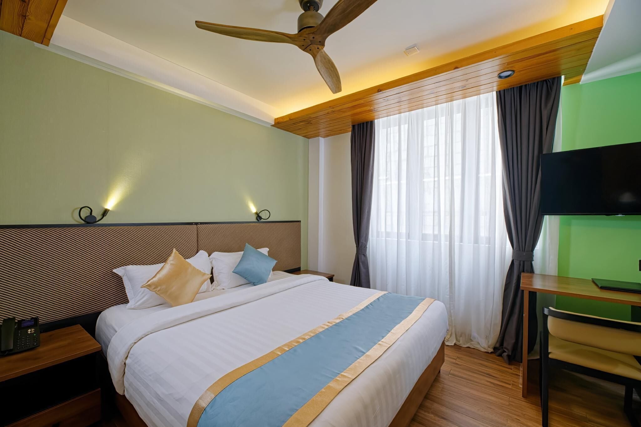 Habitación de hotel con cama, cabecero de madera, ventilador de techo, televisor y escritorio con silla.