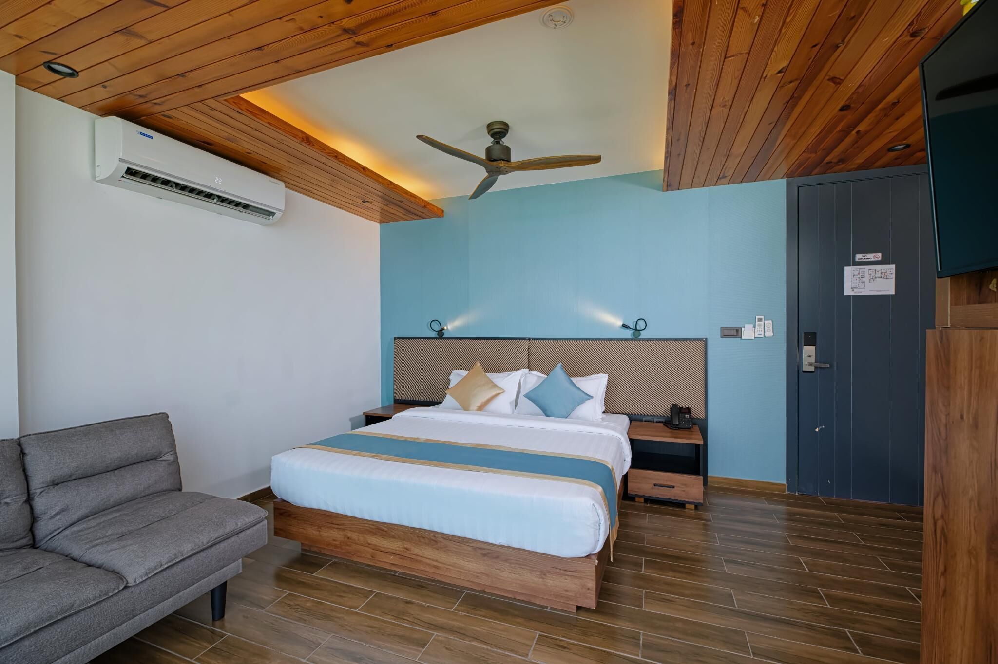 A hotel room with a bed, a sofa, a ceiling fan, an air conditioner, and a door.