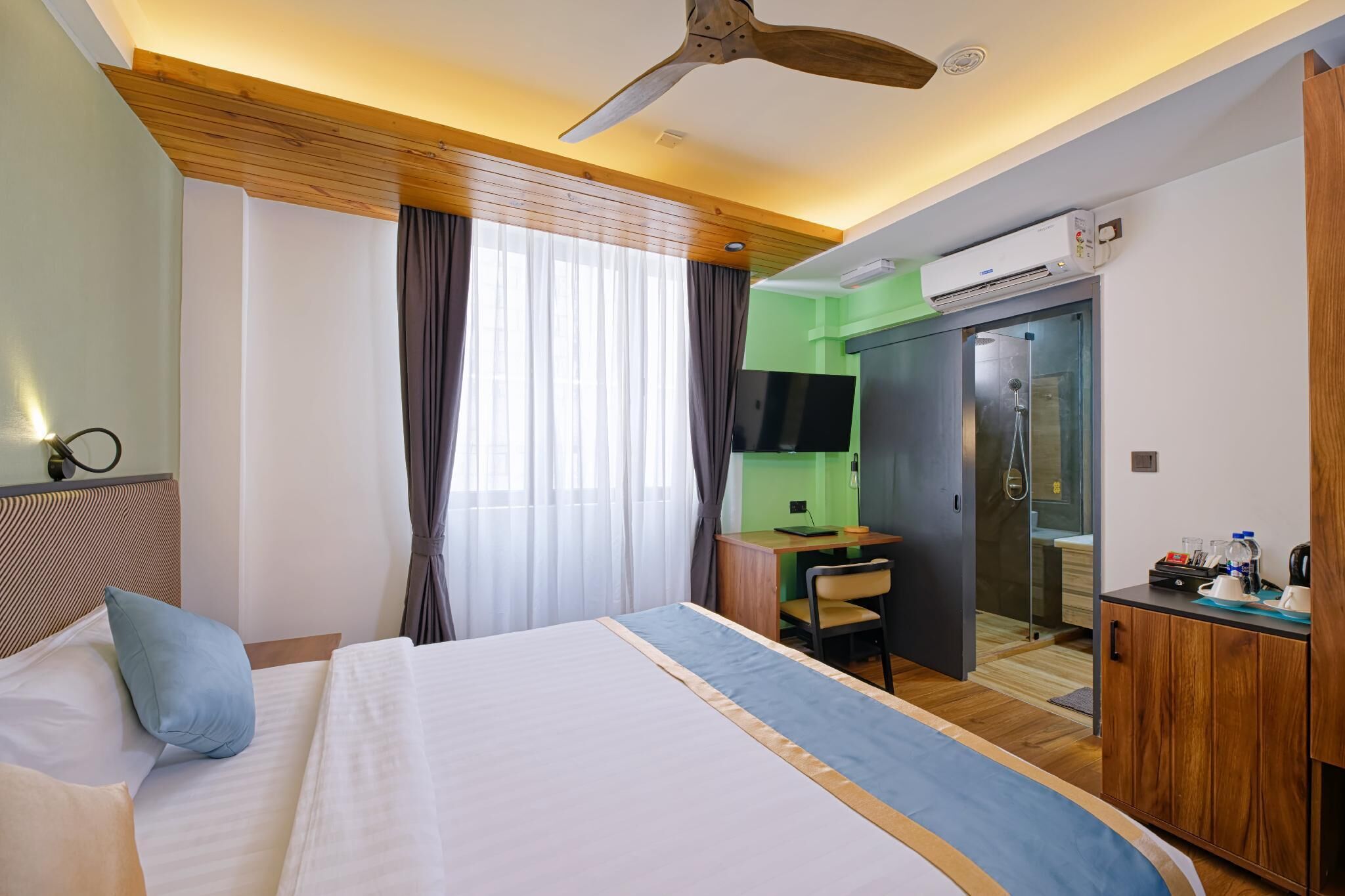 A hotel room with a bed, a desk, a chair, a wardrobe, a ceiling fan, and an air conditioning unit.