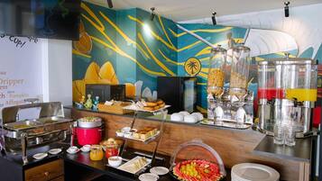 A buffet setup with various food items and beverages, including a red hot pot, a glass jar of pickles, and a fruit bowl.