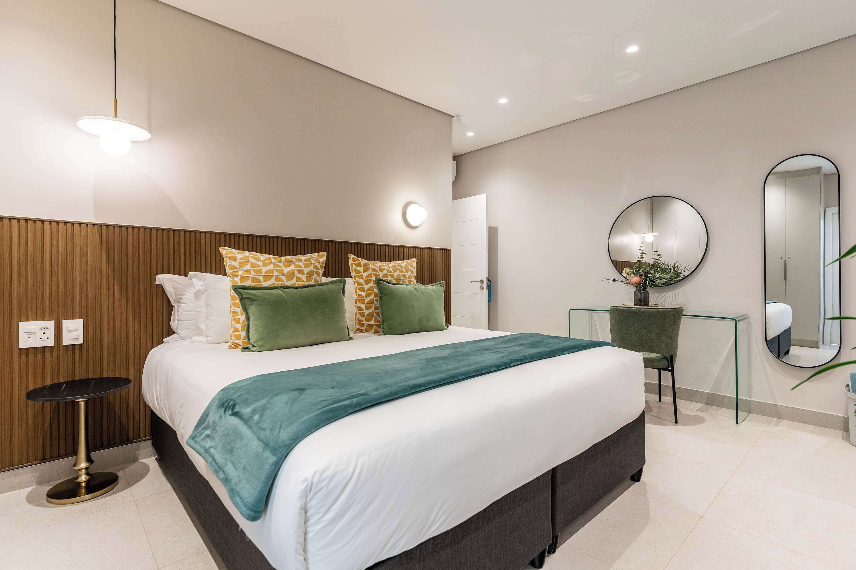 A modern hotel room with a large bed, a small desk, a round mirror, and a green chair.