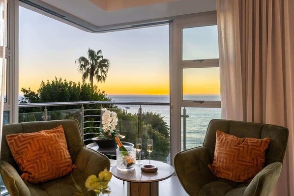 A balcony with two armchairs, a small table with a bottle and glasses, and a view of the sunset over the sea.
