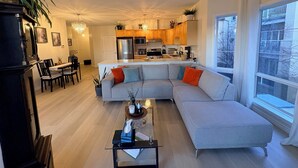 A modern living room with a large L-shaped sofa, a dining area with a table and chairs, and a kitchen with wooden cabinets and stainless steel appliances.