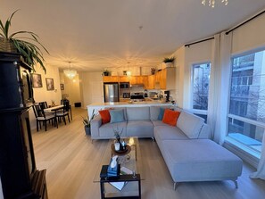 Un salon moderne doté d’un grand canapé en forme de L, d’un coin repas avec une table et des chaises, et d’une cuisine avec des meubles en bois et des appareils en acier inoxydable.